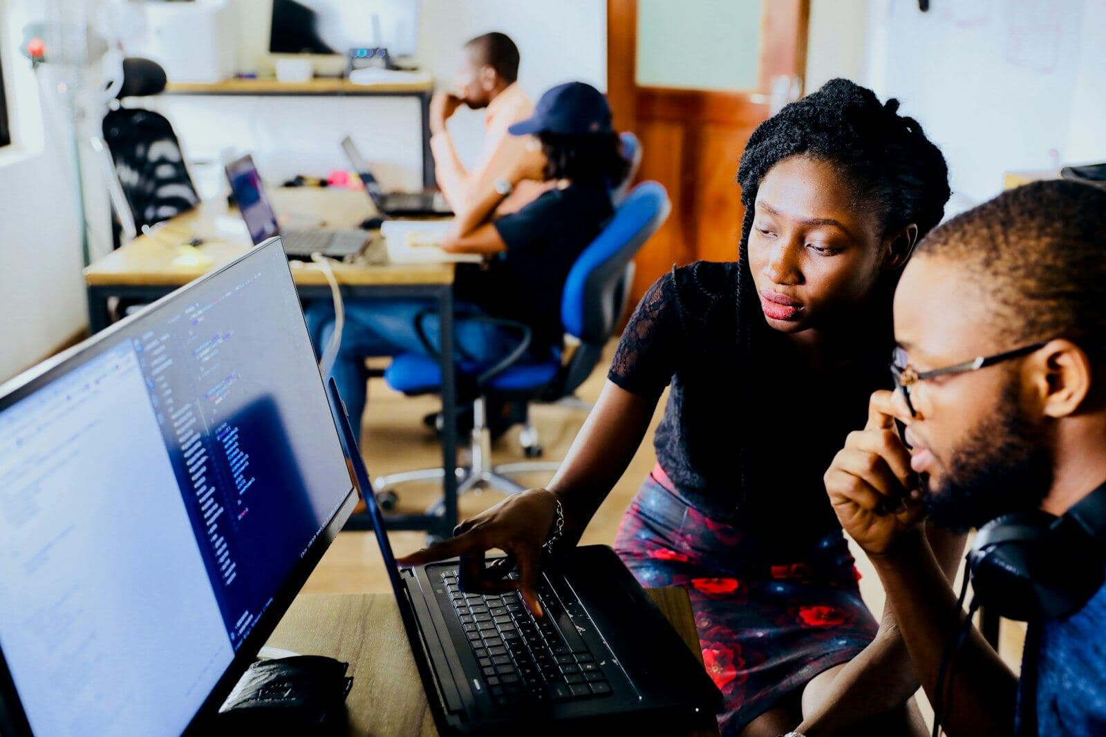 Two colleagues working through a task on a laptop, with the rest of the office at their desks